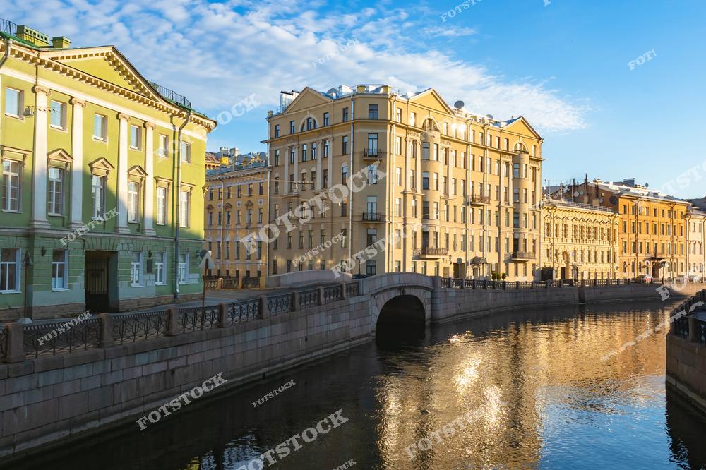 На фотографии изображена набережная реки Мойки в Санкт-Петербурге. Здания в классическом стиле освещены мягким утренним светом, их отражение заметно в спокойной глади воды. Каменные мосты и изящные ограды подчеркивают гармонию архитектуры и природы. Это место является частью исторического центра города, привлекающего жителей и туристов.